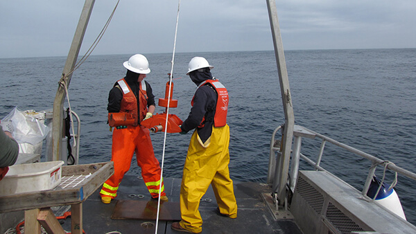 Two people in foul weather gear and hard hats stand on the back of a vessel at sea holding an orange-colored scientific instrument that is hanging off an A-frame hoist.