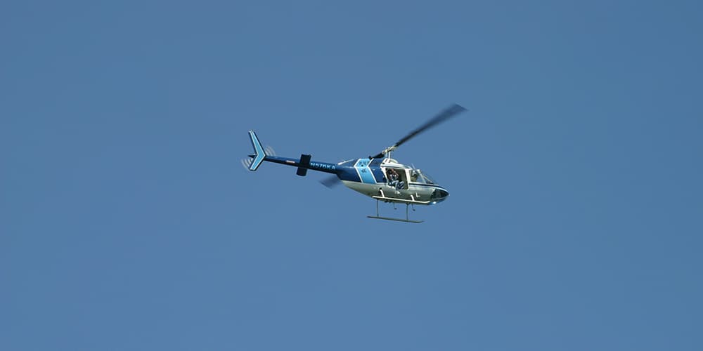 A blue helicopter, with a lighter blue stripe on the back of the cabin and on its tail, is in flight.