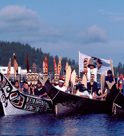 Multiple tribal canoes raft together filled with paddlers who hold their paddles up while practicing protocol on arrival. A tribal flag is flown on one of the canoes. 