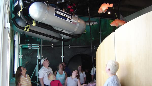 A family stands inside a visitor center looking at a model of a submersible research vessel that is on display along with various fish in the background