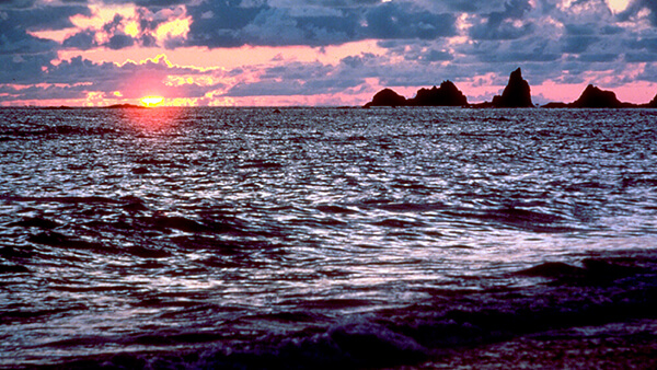 The sky is red and purple as the sun sets on the ocean with several sea stacks on the horizon.