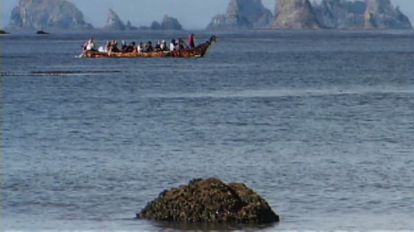 A tribal canoe with multiple paddlers is making its way towards the shore with rocky islands seen in the distance