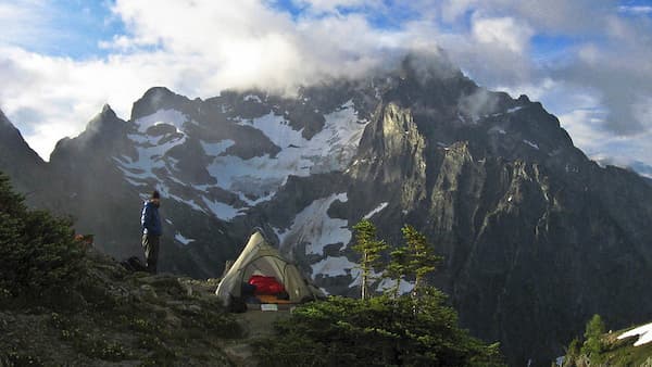 A camper stands next to their tent on a remote forested summit, overlooking a snowy mountain