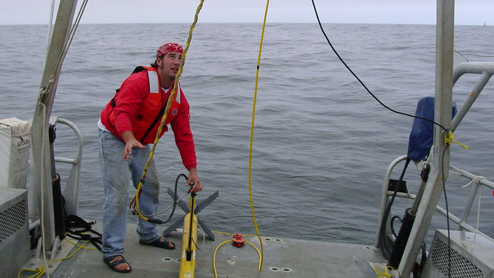 At sea, a scientist on the back of a boat pays attention to a hanging cable that is attached to a yellow torpedo-looking device that is a sonar. 