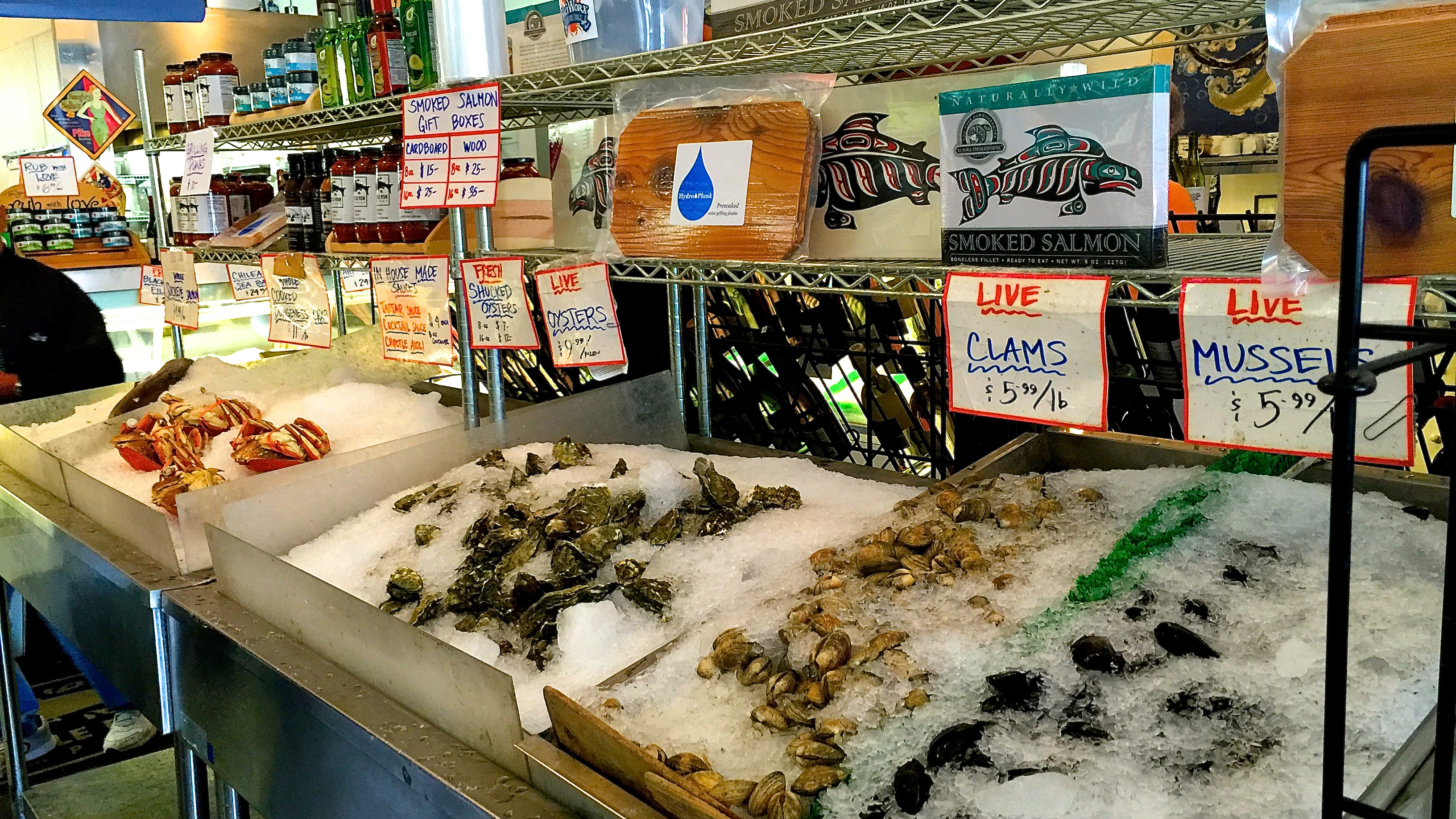 A fish market showcases fresh crab, oysters, clams, and mussels on beds of ice, along with boxes of Native American wild caught salmon on the shelves above.