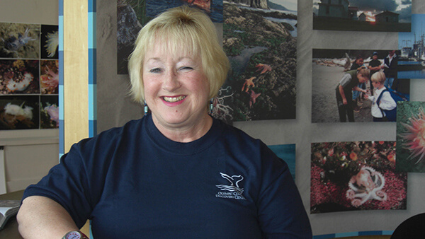 A smiling woman wearing a sanctuary t-shirt sits in front of a display showing images of scenes and activities in Olympic Coast National Marine Sanctuary