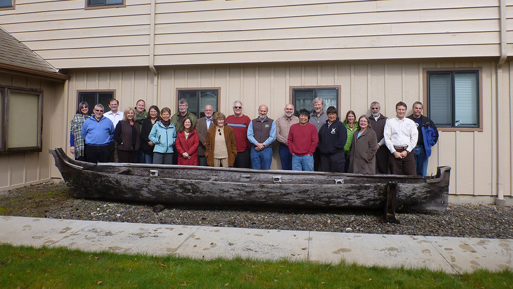 A group of people stand together in front of a beige building. A wooden canoe is in front of them on the ground