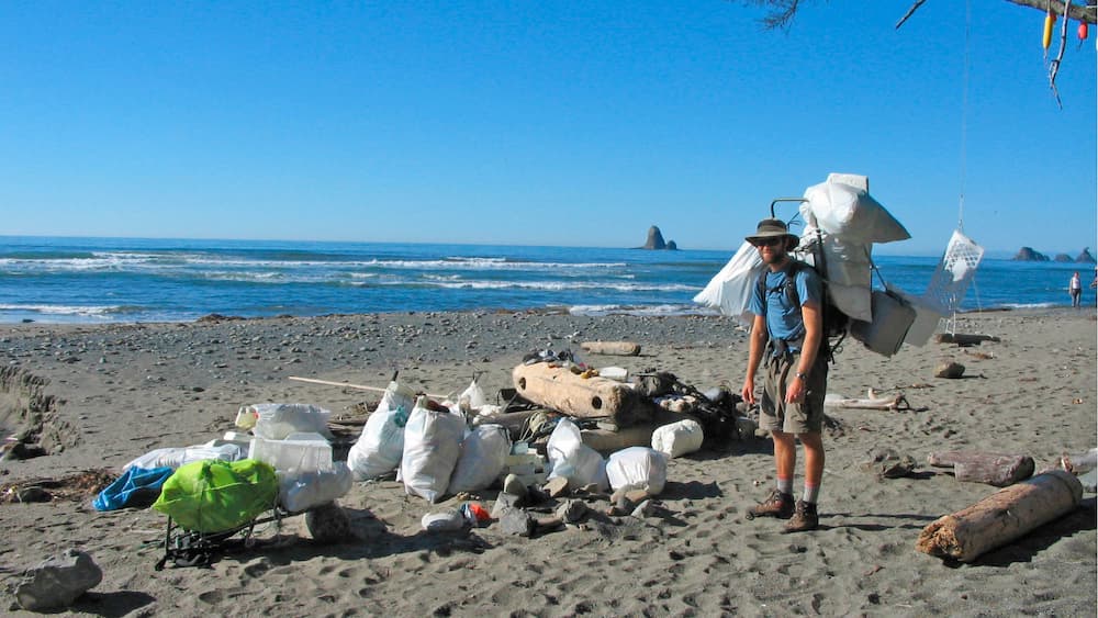 A man with a large framed backpack filled with trash stands on the beach next to a pile of filled trash bags and other marine debris. In the background there are ocean waves, as well as sea stacks in the distance.