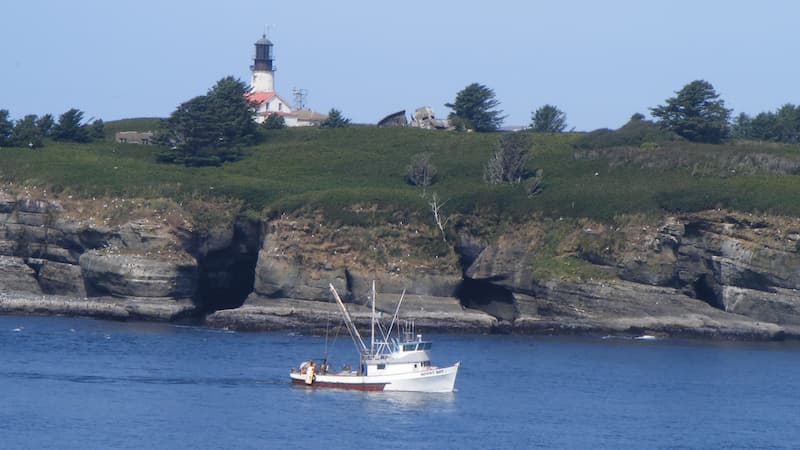 A fishing boat is underway. A rugged coastline and lighthouse is in the background.