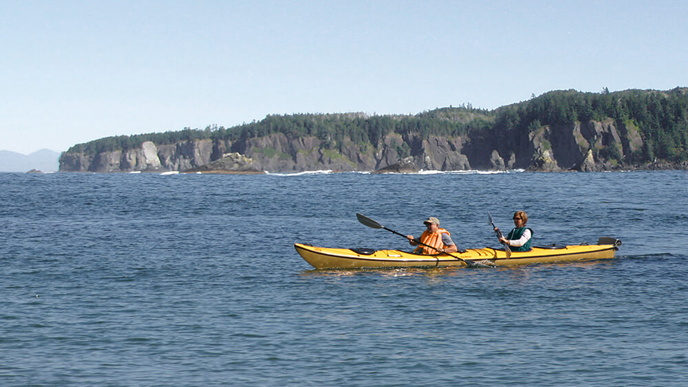 Two kayakers in lifejackets paddle in waters off a rocky coastline