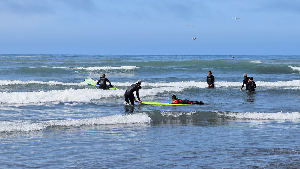 Multiple surfers prepare for waves while standing or sitting on their board in the surf.