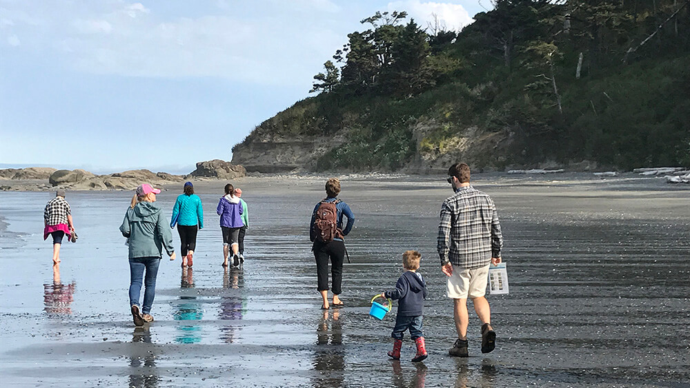 A group of people, including a small child, walk on a sandy beach at low tide towards the rocky intertidal area.