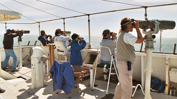 Six people use binoculars or telescopes to look off the bow of a boat at something in the water.